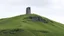 Placeholder: Solitary tower emerging from a hillside covered in green grass, contrasting with the eroded rock at the top.