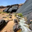 Placeholder: Fotografia di un paesaggio desertico con ruscello con polvere simile a malta e calcina, piante, rocce ruvide scure e corrose dallo scorrere dell'acqua, parete di roccia nera levigata e in parte ruvida con aperture