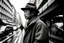 Placeholder: fotografía en blanco y negro, un hombre vestido con abrigo y sombrero de ala ancha, en un calle de una ciudad japonesa, figura difuminada en primer plano, mirada lateral, de fondo las líneas de la perspectiva se entrelazan uniéndose en un punto al fondo, arrecia una tormenta, fotografía realizada con cámara Leica y objetivo de 35 mm