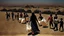 Placeholder: A Palestinian woman wearing a dress carrying very large bags of flour on her back, bending her back down in the destroyed Gaza City, and aid boxes descending from planes near the sea, with a large number of children looking up.