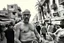 Placeholder: man looking like "Walter White" in breaking bad tv show, bald and goatee glasses, with bare chest riding on a camel in a alley Casablanca town, panorama, black and white photo, lot of markets and people in background