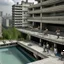 Placeholder: Several people of different genders, ages and races participate in different activities on the balconies and ledges of a brutalist concrete building with a swimming pool at the bottom, overlooking a dense urban city