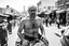 Placeholder: man looking like "Walter White" in breaking bad tv show, bald and goatee glasses, with bare chest riding on a camel in a alley Casablanca town, panorama, black and white photo, lot of markets and people in background