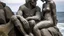 Placeholder: Two figures, female and male, are joined and embraced. Carved from textured, weathered stone with visible cracks, they resemble humanoids fused with shark heads, seated on a rocky outcrop overlooking a turbulent sea. In the background, a ghostly sailboat.