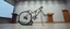 Placeholder: pinhole cam, airbrush 3d rendering, on tv screen:A five-second cinematic single take in a clean industrial presentation hall. A bizarre mechanically plausible tracked bicycle machine stands beside a presentation podium, designed to look like a giant mechanical praying mantis. Two large bicycle wheels form its main body and four thin articulated mechanical legs support it. Two long folded mechanical front arms resemble mantis forelegs