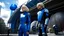 Placeholder: A full-body photograph of a young, slender woman with blond shoulder-length bob hair in a blue jumpsuit, standing in a futuristic cityscape with advanced spacecraft behind them, with a man in a huge metal exosuit with a helmet standing guard beside her.