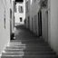 Placeholder: Street of a town on an Italian island in summer, with stairs and arches, decadent tone, real photography, photography taken with a Leica camera and 50 mm lens, following the style of the 'Ripley' series, black and white photography, toned 50s tones