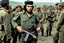 Placeholder: color image of Che Guevara with his real face and hat with star standing with heavy gun in hand boots in a fighting area, other soldiers are fighting