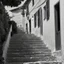 Placeholder: Calle de un pueblo de una isla italiana en verano, con escalinata, fotografía real, fotografía realizada con cámara Leica y objetivo de 50 mm, siguiendo estilo de la serie 'Ripley' emitida en Netflix, fotografía en blanco y negro, virada tonos años 50