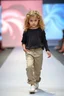 Placeholder: 2 years old beautiful baby blue eyes girl fashion model in loose pants and loose top with blonde curvy wavy hair ,cat walks in runway,catwalk and pose at end with cute