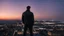 Placeholder: An Englishman in a bomber jacket standing to one side of a tall building looking across a city after sunset