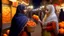 Placeholder: A full-length Palestinian girl wearing an embroidered dress and a white embroidered shawl buys oranges from an old seller wearing a keffiyeh in the market of Jerusalem, 100 years ago, at night with multi-colored lights reflecting on her.