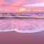 Placeholder: a serene and picturesque seascape, likely captured during either sunrise or sunset. The scene captures a tranquil moment by the sea, with the sky illuminated by warm colors ranging from soft pinks to deep purples. These colors reflect beautifully on the water and wet sand below. The gentle waves softly crash on the shore, creating a frothy white surf that contrasts elegantly with the pink-tinted water and sand. In the distance, fluffy clouds in shades of pink and purple are scattered across the