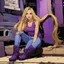 Placeholder: A girl with a purple gold hue around them with small bits of purple gold on their skin. They have long, dirty blonde hair and wear a tank-top with a jacket around their waist and jeans. They wear boots and have violate eyes.