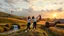 Placeholder: a group of young ladies in sports pants and blouse are dancing to camera in village over high grassy hills,a small fall and river and wild flowers at river sides, trees houses ,next to Ripe wheat ready for harvest farm,windmill ,a pretty train is arriving to station,cloudy sun set sky