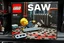 Placeholder: Lego box displayed for sale on a store shelf. Text: "SAW" as exact title on box across top in bold clear font. Extremely satirical dark scene showing Lego blonde haired male minifigure laying on the floor chained by leg to detailed Lego radiator in grimy abandoned boiler room. Pronounced raised pip textures on all Lego surfaces. Focus on gray Lego hacksaw on floor in foreground, just within minifigure's reach. Yellow minifigure with terrified expression. Rusty pipes and dripping water effects bu