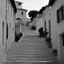 Placeholder: Calle de un pueblo de una isla italiana en verano, con escalinata, y arcos, tono decadente, fotografía real, fotografía realizada con cámara Leica y objetivo de 50 mm, siguiendo estilo de la serie 'Ripley', fotografía en blanco y negro, virada tonos años 50