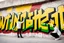 Placeholder: painted on concrete wall of underpass a young cyberpunk hacker paints a complicated colorful / uppercase all-white graffiti text in text: "WINTERMUTE"; street art, graffiti green-yellow-black triotone, wall painted matte black and yellow and deep red, semi-front view, low angle, daylight, cinematic, dramatic