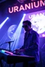 Placeholder: French cool synth player, he is called "Jean-Maurice Brûlée" performing at dance club, his name is "Jean-Maurice Brûlée" , white and blue Neon lights and smog, an earth is projected in background, and "URANIUM" big sign on the wall, he is playing on synthesizers on stage with lot of laser beams and wearing black sunglasses,