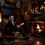 Placeholder: A pleasant and charming scene of a 70-year-old man in the Victorian period sitting on a wing chair next to the fireplace and reading The Times of London while enjoying a glass of wine on a winter evening. The man has a balding forehead and curly white hair. He wears black shoes and sits by the hearth on which candles are lit in candlesticks. On the wall hang paintings of an English countryside landscape. A boxer dog is napping on a carpet on the floor next to the woman. . The room is lit by th