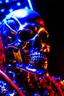 Placeholder: A close up of a skeleton face looking at the camera from a mysterious side view. Deep bony features and inside the hollow eyes are red shining lights, scary. Dressed in an astronaut suit floating in space. On his suit is an American flag and in his one hand is a small wavering American hand flag. From the back of his suit is blowing out blue, white and red smoke. Realistic, 8k, highly detailed, funny