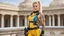 Placeholder: A happy modern Barbarella warrior with a machine gun covered by tattoos, 24-years-old her lean muscular frame wrapped in a yellow tactical bodysuit and a bullet proof vest over her bossom. Her blonde hair is pulled back in a tight ponytail, revealing blue eyes, she stands on the balcony of the marble palace of a modern dictator, behind we see the beach and palm trees [vintage style noisy scratches glitches, Shot the texture and then rewound the film and double exposed from a 1970 B series movie]