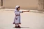 Placeholder: A five-year-old Palestinian girl wearing a traditional dress and new shoes looks to the side and points at a distant building.