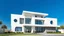 Placeholder: Summary: The image depicts a modern architectural building set against a clear blue sky, featuring sleek lines and circular windows.Detailed Description:Objects: The main object is a contemporary house with a combination of flat and curved structural elements. The building has large glass windows that suggest an open and airy interior, while the exterior features a mix of smooth white and gray surfaces.People: Two individuals are visible in the scene, positioned near the entrance, engaging with