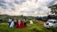 Placeholder: group of people are dancing in a national celebration in a village over high grassy hills,a small fall and river and wild flowers at river sides, trees houses ,next to Ripe wheat ready for harvest farm,windmill ,a few village local shops .people are dancing in a wedding celebration,a pretty car with flowers for eedding couple, cloudy sun set sky,a few village local shops