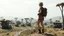 Placeholder: A photorealistic image of a slender 60-year-old woman, in brown shorts, a shirt and boots, with a backpack, standing on a hill looking down on a derelict sci-fi village with low alien buildings with trees and fungi in clumps.