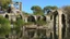 Placeholder: A ruined stone building partially submerged in a lake, balconies, verandas, arches, bridges, spires, stairs, trees, dense foliage, spanish moss, ivy, blue sky, white clouds