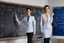 Placeholder: A pretty brown-haired, light-skinned woman in an elegant blue suit stands in a classroom in front of a blackboard with various charts and diagrams, in the sunlight