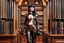 Placeholder: a full-length pale dark-haired woman with a straight bob hairstyle with a fringe, in a steampunk leather outfit, and gloves, standing in a steampunk library