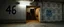 Placeholder: background of wall(wet textured concrete, gray, old, cracked, stained, 1 wall light) from underground parking, hong kong style, a large number "46" painted on left(old faded paint), at middle is an opening into an area with stairway(poorly lit), cyberpunk, to the right of opening is a faded rectangle