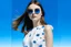 Placeholder: On a pop sky blue background, with embedded daisy flowers, a woman in a white dress, brunette, round sunglasses, 16K, real photography, advertising photography