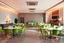 Placeholder: A 3D hyperrealistic image in 4K resolution of a modern party and birthday room approximately 100 square meters in size, with elegantly arranged tables, vibrant green chairs, and an abundance of warm soft light casting a celebratory ambiance, featuring a sleek kitchenette with stainless steel appliances and polished countertops, where guests can gather and mingle, surrounded by the warm glow of tabletop lamps and the subtle sheen of glossy flooring, with every detail meticulously rendered to crea