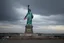Placeholder: the statue of liberty, dress dirty and torn, her face solemn and terse, trash littered at her feet, she holds a crumpled american flag, she walks away with back turned under a dark and dreary sky, wide angle