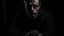 Placeholder: In the photo, we see a man sitting in the dark, looking at the camera with a cold gaze. The man is dressed in black and seems uninterested in what is going on around him.