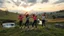 Placeholder: a group of young ladies in sports pants and blouse are dancing to camera in village over high grassy hills,a small fall and river and wild flowers at river sides, trees houses ,next to wheat farm,cloudy sun set sky