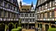 Placeholder: A Paved Courtyard, With Tudor Gothic Houses, Tall twisted Chimneys, warped Rooves, People, Shops,