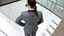 Placeholder: In a high-angle perspective, a woman is seen standing in front of a staircase, her back to the viewer. She is dressed in a gray suit jacket, gray pants, and a black collared shirt. Her hair is neatly pulled back into a ponytail and she is adorned with a pair of black sunglasses. The staircase, built of white marble, has a glass railing on each side. To the left of the staircase, a brown-framed window adds a touch of natural light. The light brown wooden floor is adorned with a few cushions on bo