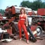 Placeholder: [Mid 1980s Magnetism and chainsaw from my personal archive scanned from original 35mm slide] glamour shot of Wincey Willis : she is impressive, she stands in the junkyard a chainsaw in hand, in her red revealing swimsuit