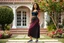 Placeholder: very beautiful lady with beautiful midi dark black-red skirt and top with curvy hair sports shoes standing in a nice villa yard with trees flowers ,16 by 9