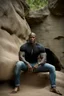 Placeholder: Alimi Ballard as very muscular male. bald with tribal tattoos wearing jeans and a teeshirt outside a cave