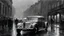 Placeholder: [grayscale : vivid: 16] painting depicting a silver (elongated:1.3) 1934 Rolls Royce Phantom II Continental on the streets of 1930's London, rear view, rainstorm, visible rain, dark sky, man holding umbrella in background, highly detailed, absurdres, masterpiece,