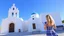 Placeholder: Picturesque scene of a traditional Greek Orthodox church with a white facade and a striking blue dome, set against a clear blue sky. The church features a bell tower with multiple arches and bells, and a wooden door adorned with intricate carvings. A woman with light skin and blonde hair, wearing a blue and white dress, is walking away from the camera towards the church, holding a selfie stick. The ground is paved with light-colored stones, and there are a few potted plants and a bench on the le