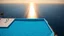 Placeholder: majestic infinity pool merging seamlessly with the ocean horizon, impossibly crystal blue water surface reflecting the sky like a perfect mirror, creating the illusion of swimming between two atmospheres, minimalist white marble platform appearing to float above the cliff, tiny human figures at the edge adding dramatic scale to the immensity, sunbeams piercing the water creating hypnotic light patterns in the background, solitary palm trees casting elongated shadows at sunset, ultra-modern archi