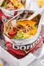 Placeholder: "Taco in a Bag" which consists of an open Doritos chip bag with sides rolled down, containing Doritos chips and cooked ground beef and lettuce and shredded cheese and chopped tomato pepper and onions and topped with more nacho chips, food blogger photography