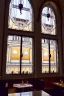 Placeholder: View of the synagogue in Budapest from a large glass window of a café, a brown-haired woman sitting in front of the window with her back to us, coffee in front of her, large shiny bright silver and spherical lamps on the ceiling, in sunshine