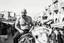 Placeholder: man looking like "Walter White" in breaking bad tv show, bald and goatee, with bare chest riding on a camel in a alley Casablanca town, panorama, black and white photo, lot of markets and people in background